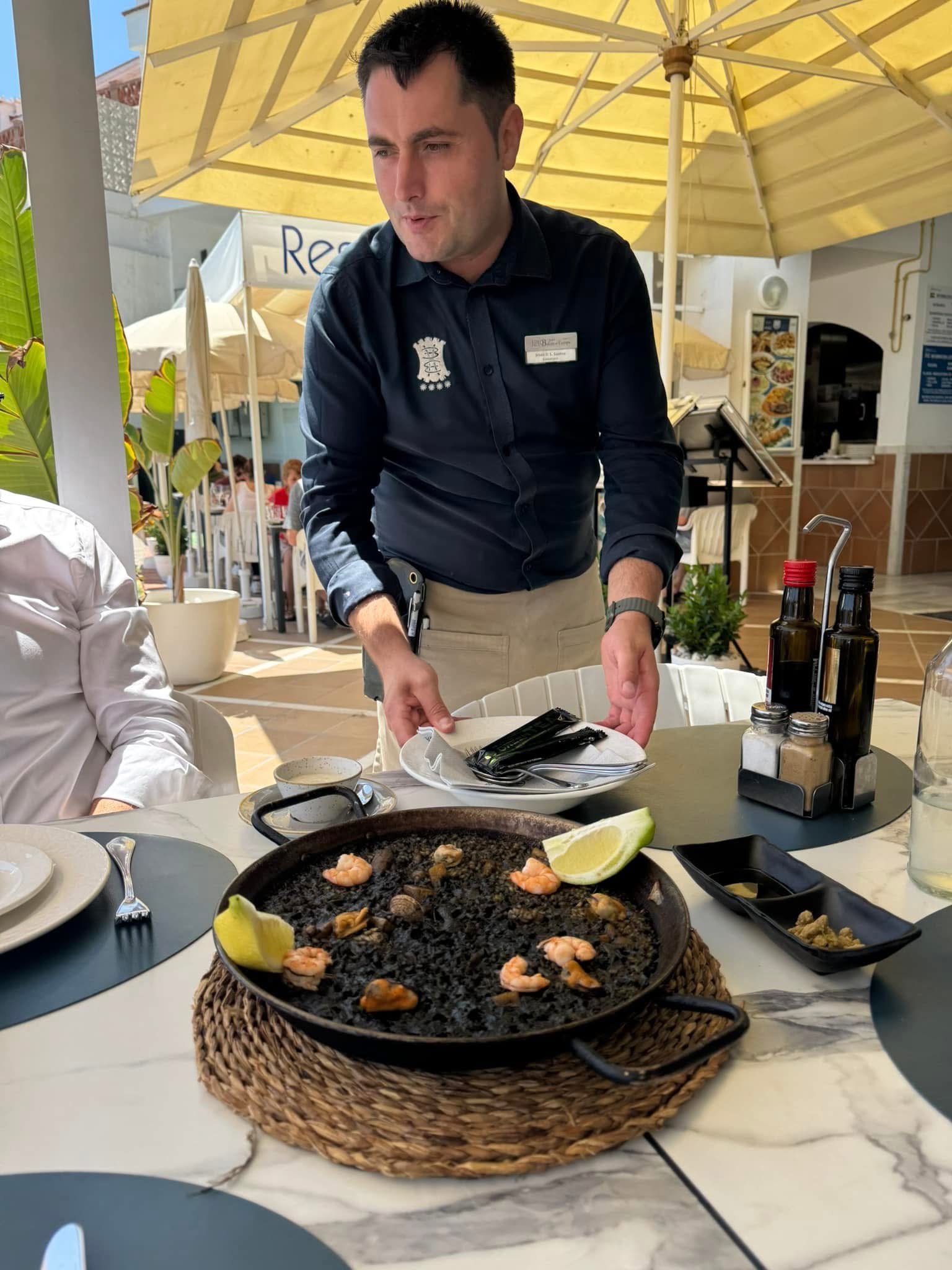 A waiter serves black seafood paella on a terrace.